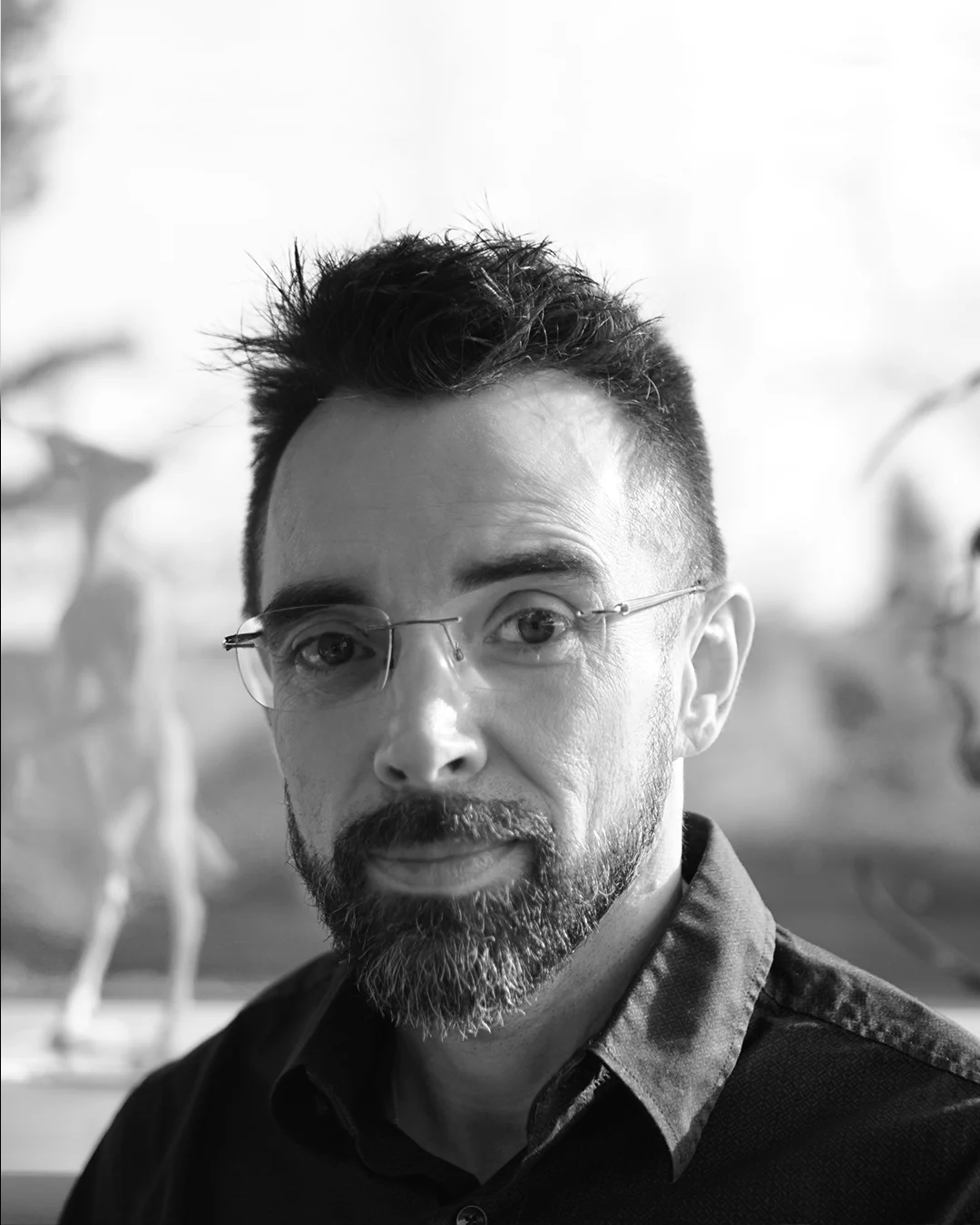 Man with glasses, beard, and shirt standing against a blurred background, likely a musician