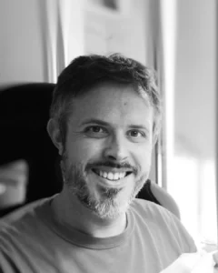 Man with a beard smiling at the camera in a black-and-white photograph, wearing a t-shirt, against a curtain backdrop