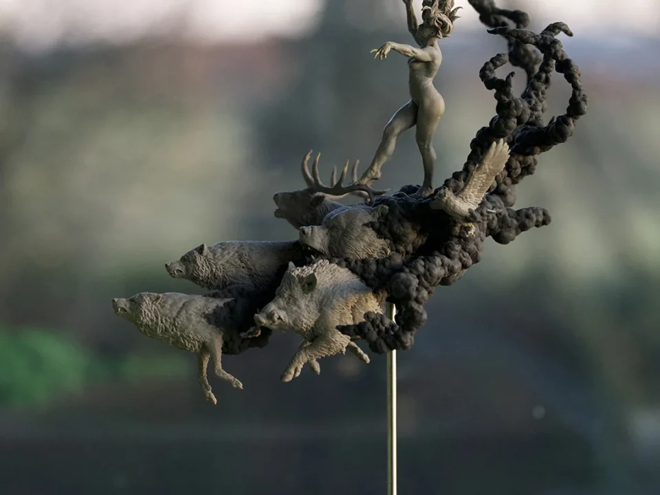 Bronze statue of a woman standing on a pile of animals, with the word 'KAZAM' visible at the base