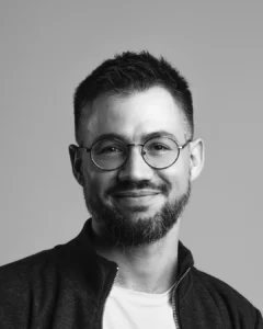 Man with glasses, beard, and jacket smiling against a plain white background