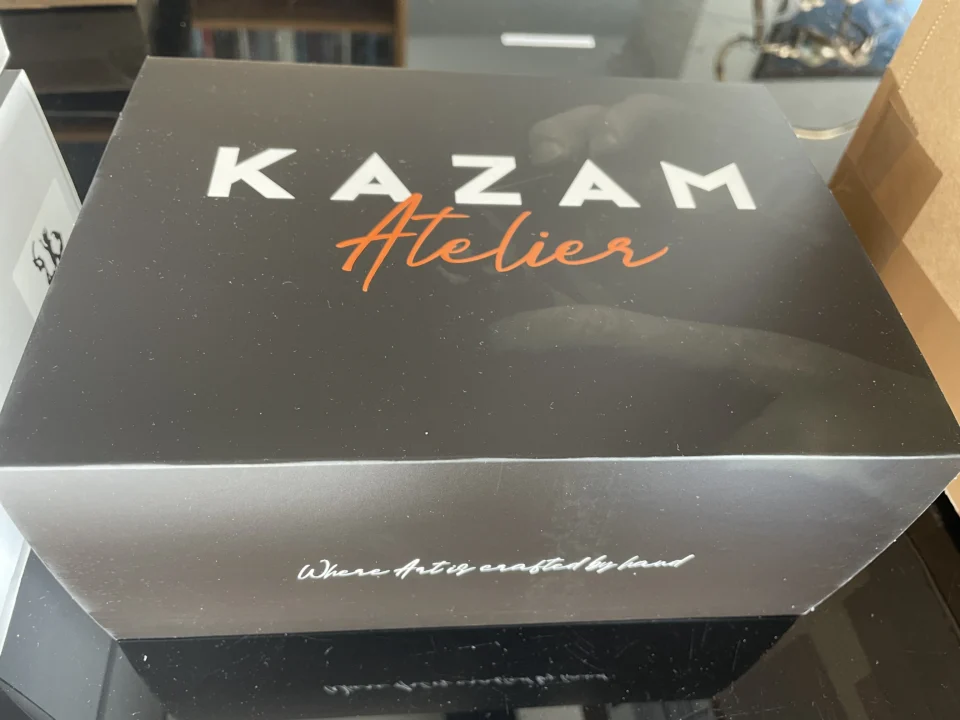 Black box with white text 'KAZAM Atelier' on a table